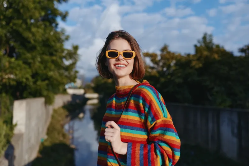 woman striped sweater yellow sunglasses smiling outdoor canal, young 20s female with short brown hair in colorful knit, casual outfit walking, joyful travel lifestyle portrait in sunlight