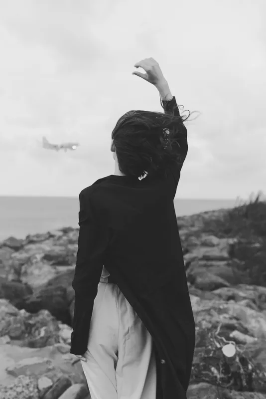 Back view of a woman on a rocky coast waving her arm toward the sky, as a distant plane passes, conveying travel, departure, freedom and mood in a moody black and white landscape
