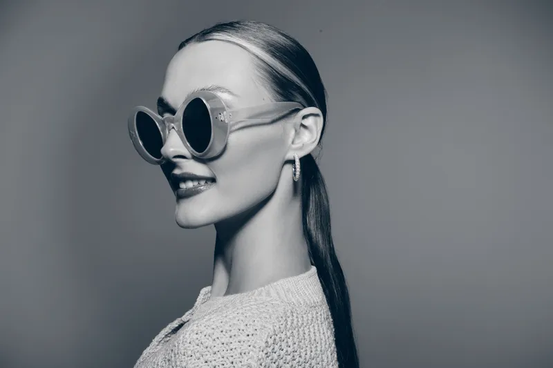Stylish Caucasian woman with pale skin wearing round sunglasses and a textured sweater looks to the side with a confident smile in a monochrome studio setting.