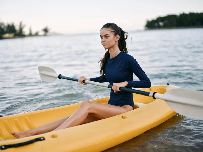 Young woman kayaking on calm water in a yellow kayak, wearing a navy blue shirt, focusing on paddling during a peaceful outdoor activity.