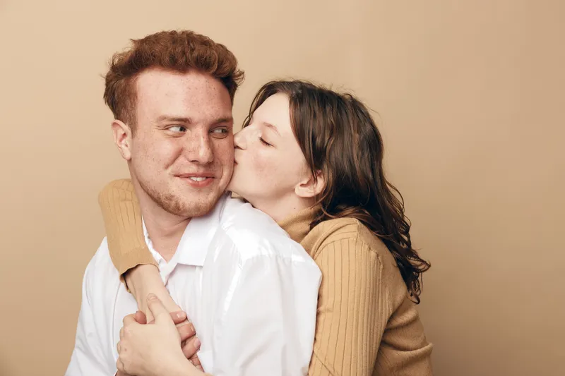 Young couple in casual clothes share affectionate moment, woman kissing man on cheek. Warm beige background creates cozy atmosphere of love, intimacy, and happiness between partners.