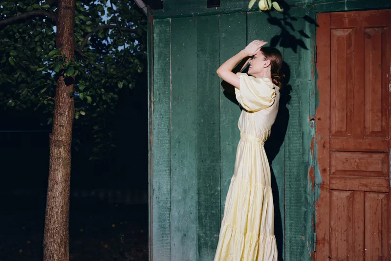 A woman in a pale yellow dress stands beside a green wall and wooden door outdoors shielding her eyes from sunlight to create a calm vintage mood and atmosphere