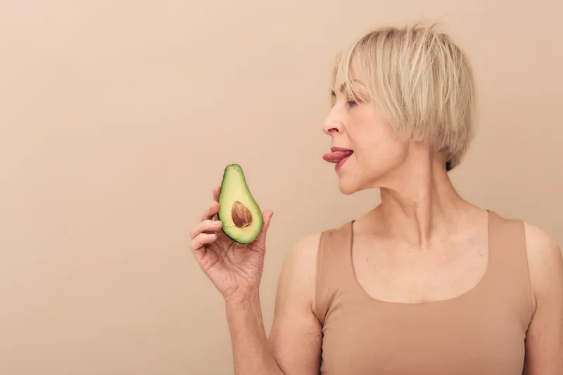 Blonde middle aged woman in beige tank top playfully sticks out tongue near fresh avocado half. Neutral background highlights natural skin texture and healthy lifestyle concept with fresh fruit.