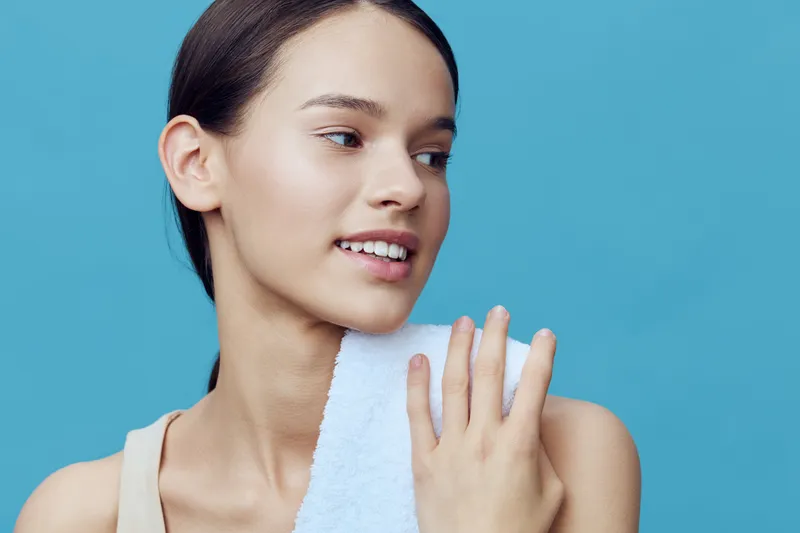 Young woman with a towel on her shoulder, smiling softly against a bright blue background, embodying health and wellness, perfect for skincare or beauty themes