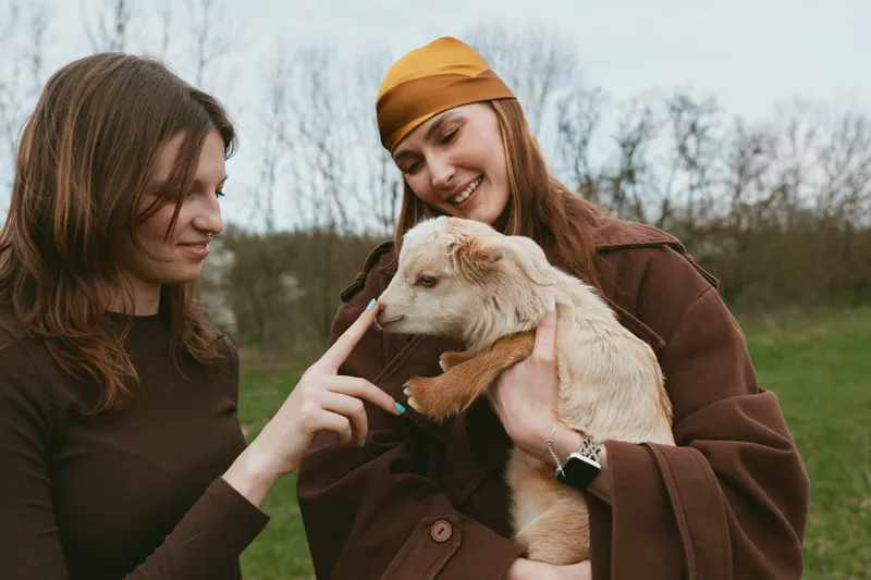 Two women outdoors with a goat kid, one gently touching the animal while the other smiles, sharing a playful moment in a grassy countryside field, rustic coats creating a warm lifestyle scene