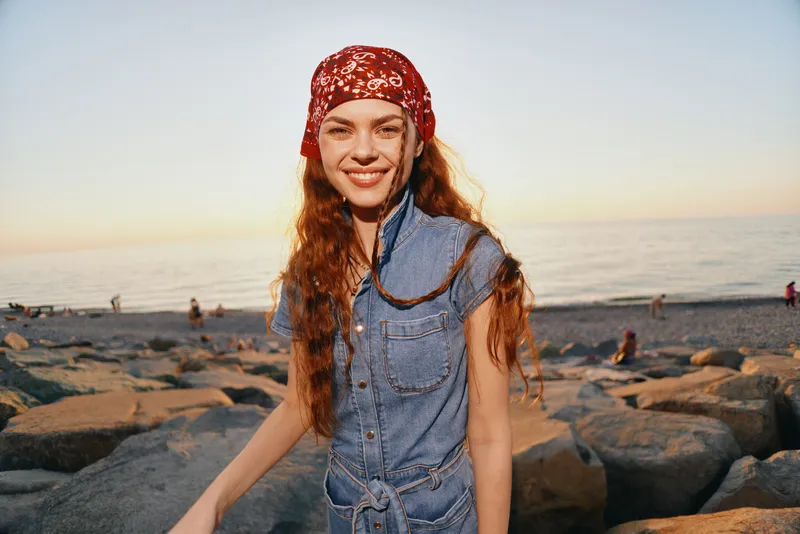 Lifestyle woman in modern boho-western denim street style smiling by the rocky beach at sunset with warm film color tones and casual summer vibe.