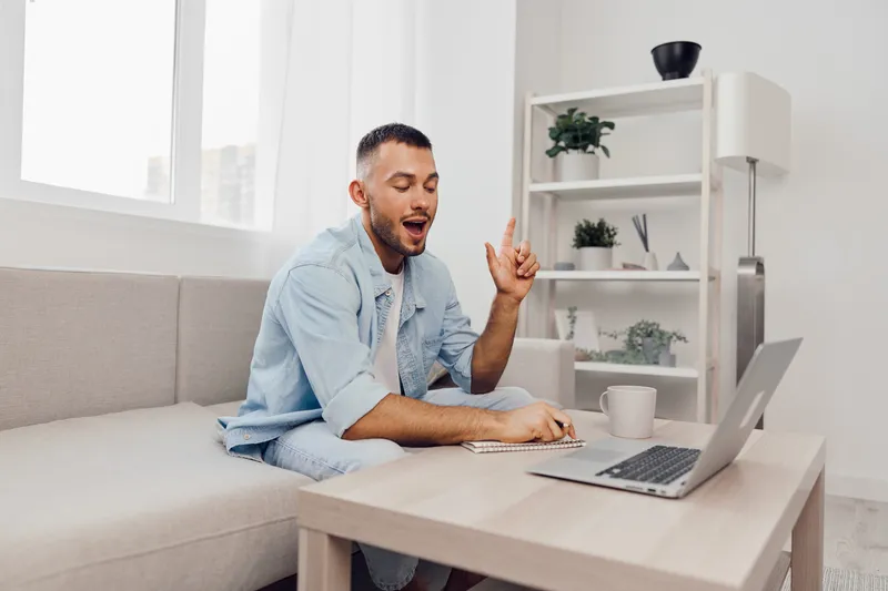 Engaging man in a light blue shirt is sitting on a sofa, working on a laptop while actively communicating, showcasing modern workspace vibes and productivity.