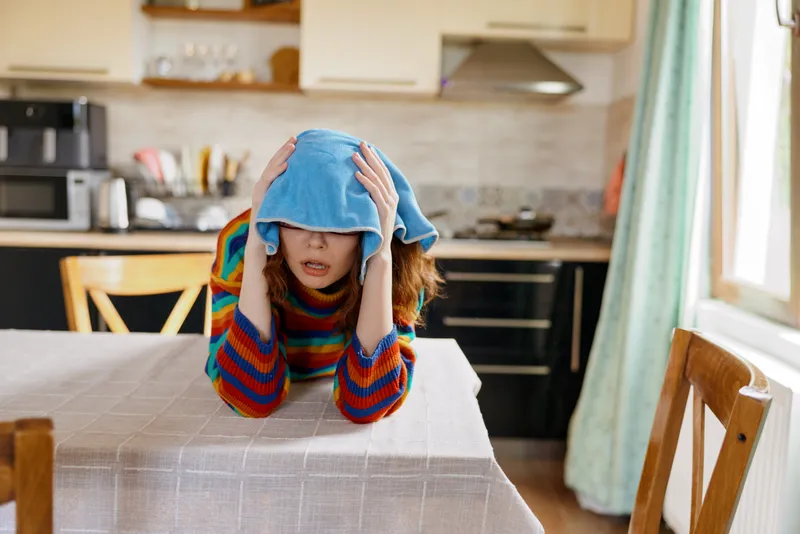 Young woman sits at a kitchen table with a blue towel over her head, looking stressed and tired after a long day.