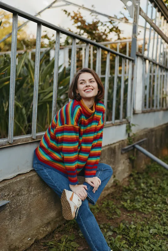 Woman smile colorful sweater jeans sitting by metal fence outdoors casual portrait of a happy young adult in a striped knit, relaxed pose with autumn greenery.