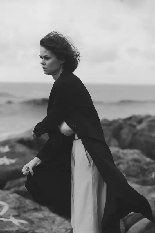 Woman, coast, coat, wind, monochrome scene of a lone figure walking along a rugged shoreline, sea spray and rocks in the background, fabric flowing with movement, moody atmosphere, stylish dramatic
