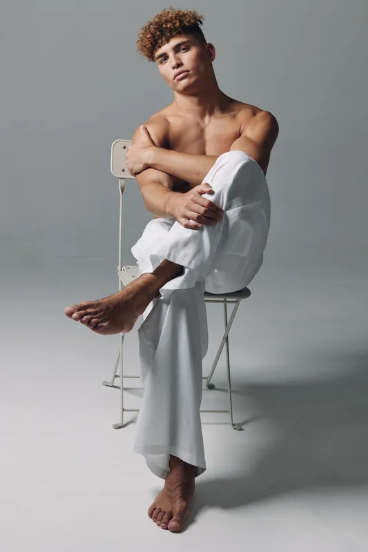 Young man with curly hair sitting barefoot on chair in white pants with crossed arms and serious expression in studio with plain background.