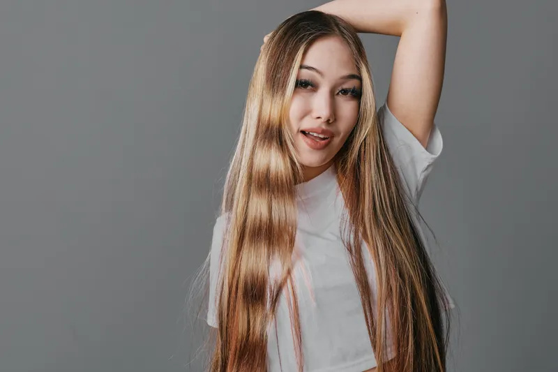 Young woman with long straight hair posing with raised arm for beauty portrait in casual white t shirt and neutral gray background indoors.