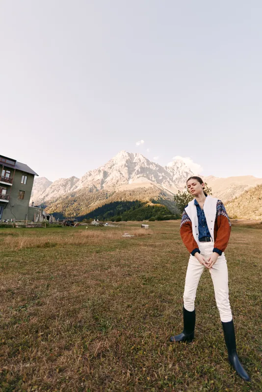 Woman model in mountains meadow wearing fashion jacket and boots, standing in outdoor landscape near a rustic building in alpine field with natural scenery and portrait pose.