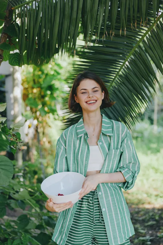 Portrait of a cheerful woman outdoors under lush palm leaves, holding a white bowl, wearing a green striped shirt and light blazer.