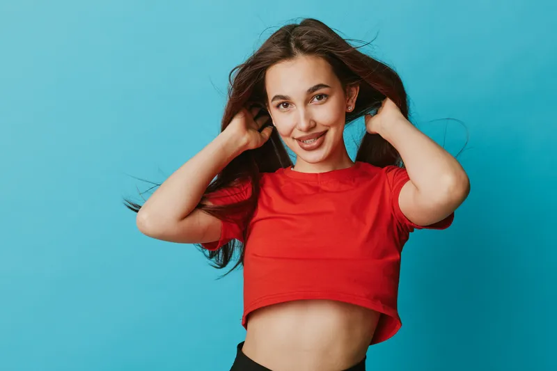 Young woman with long hair in red crop top posing with hands in hair against blue background. Smiling female model in casual clothing with natural look and confident posture.