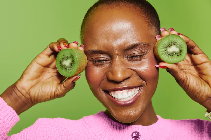 Happy African woman with dark skin smiling with closed eyes holds two halves of fresh kiwi fruit near face on green background. Concept of healthy lifestyle, fruit, and natural beauty.