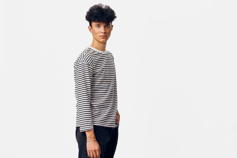 young man with curly hair wearing striped shirt and dark jeans stands confidently against white background with casual posture and thoughtful expression