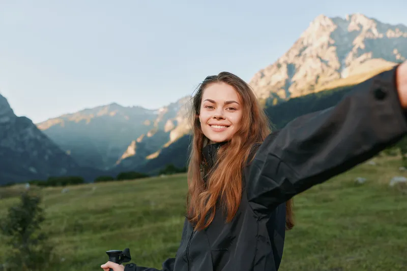 A cheerful young woman stands in a scenic mountain meadow, smiling as she takes a selfie with a compact camera, capturing a bright and energetic outdoor moment.