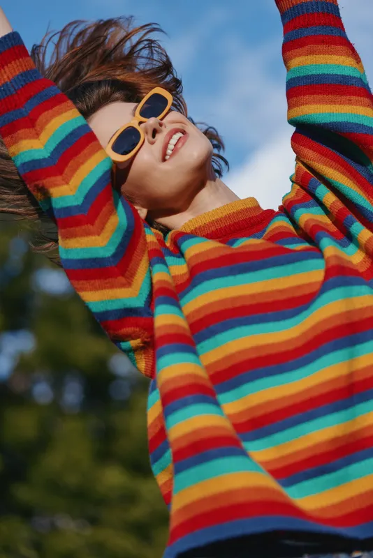 young woman in striped sweater and yellow sunglasses smiling with arms raised outdoors under blue sky, curly hair, casual outfit, joyful lifestyle portrait in a park setting.
