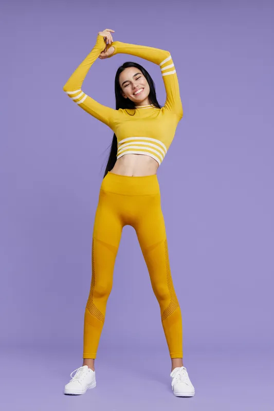 Woman sport fitness wearing yellow activewear stretching arms up confidently against purple background, showing health, energy, and well-being in workout pose.