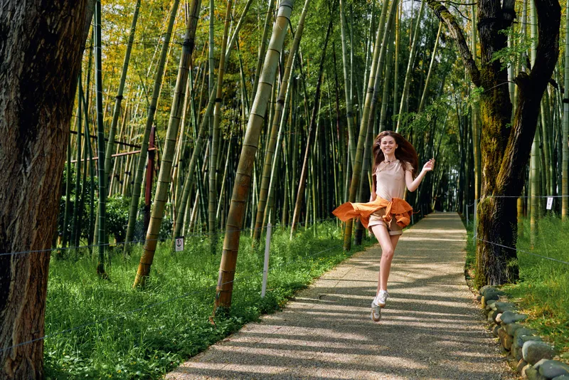 Woman rollerblades and jumps along a bamboo path in a park, smile bright as skirt twirls in motion, carefree outdoor activity on a sunny green forest trail for leisure and fitness.