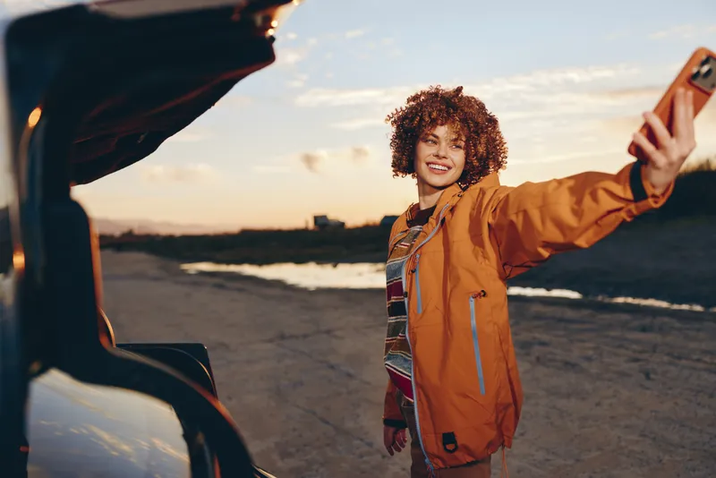 Woman lifestyle, selfie, phone held up outdoors at sunset, curly hair, orange jacket, smiling warmly near a parked car, evening beach background, joyful travel mood, casual candid moment