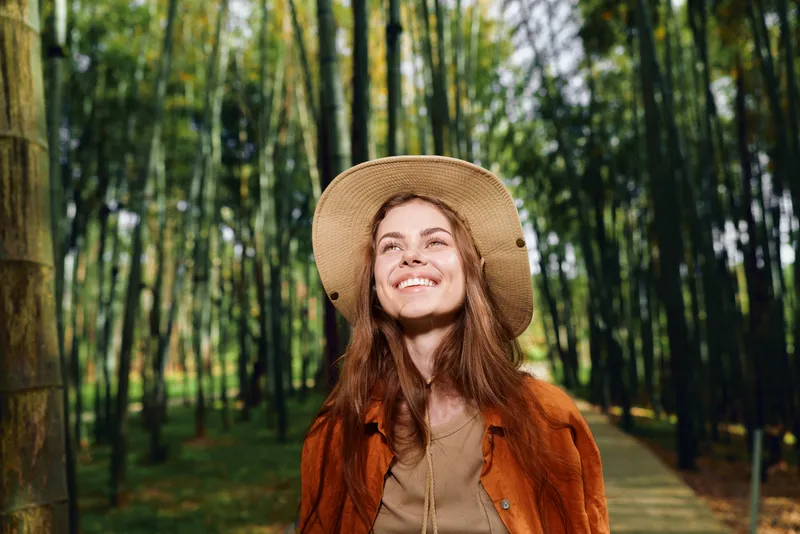 Woman smiling in a bamboo forest wearing a straw hat and brown coat on a leafy pathway, outdoor portrait capturing sunlight, happy travel mood, nature exploration and serene vibes.