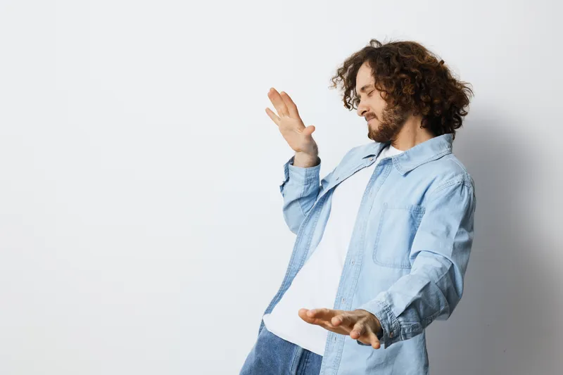 Young man with curly hair in denim shirt reacting with playful surprise against a light background, expressing joy and carefree emotions in a casual setting