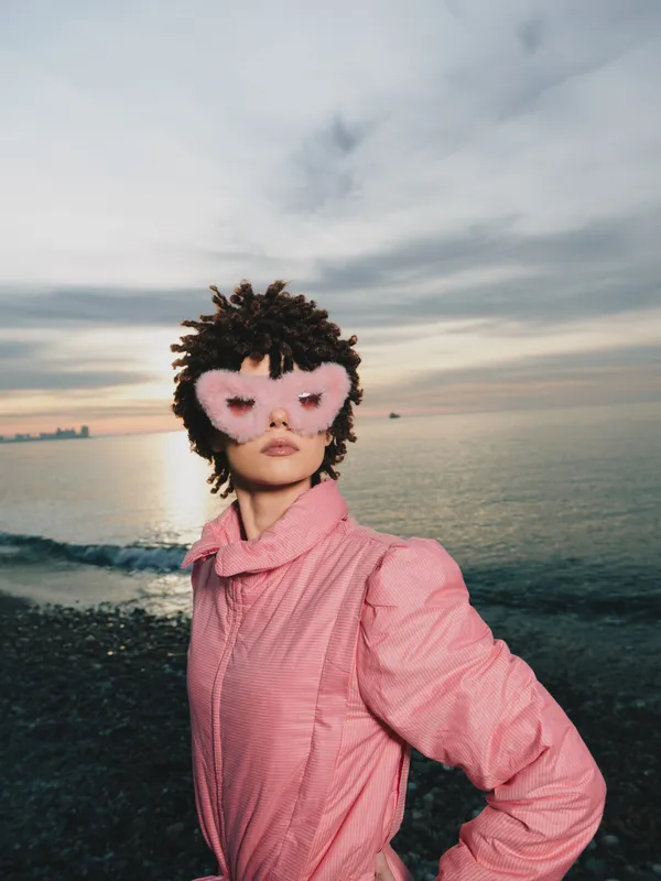 portrait on a beach at sunset featuring a person in a pink jacket wearing a playful mask, a bold fashion moment by the sea under warm light and cinematic mood