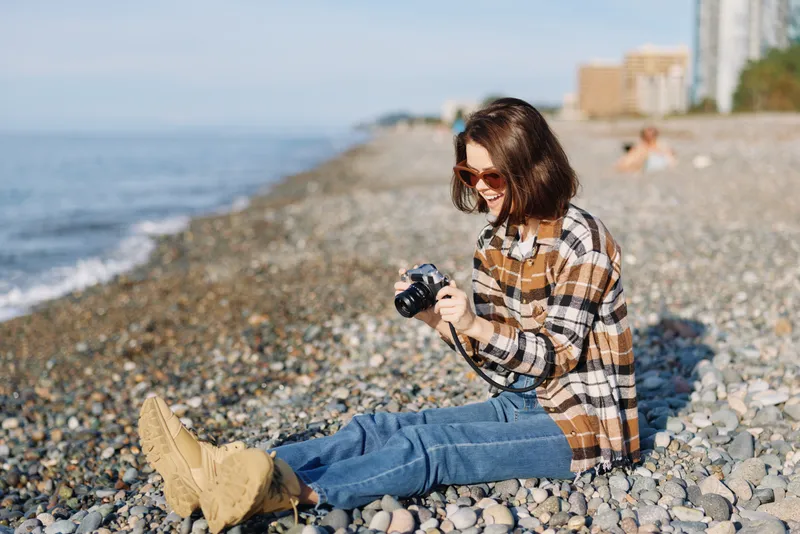 Woman on a pebbled beach with a camera, wearing plaid shirt and denim, enjoying sunny outdoor photography by the sea, with relaxed posture and sunglasses.