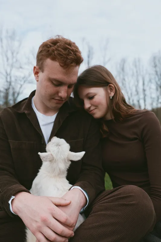 Couple sits close with a goat kid in an outdoor setting, sharing a tender moment. Warm, intimate, and natural lifestyle imagery with a sense of calm and rural charm.