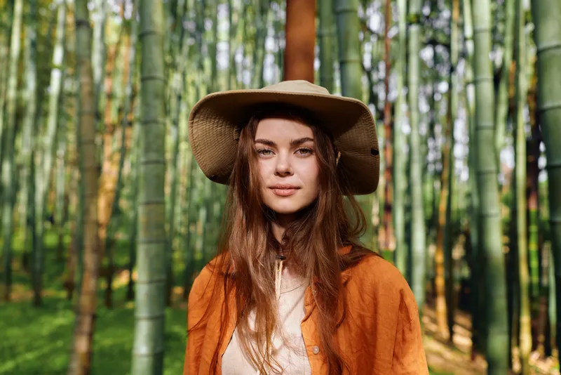 Woman portrait in bamboo forest wearing wide brim hat and orange coat, nature outdoors scene with soft sunlight, serene traveler portrait and natural background foliage.