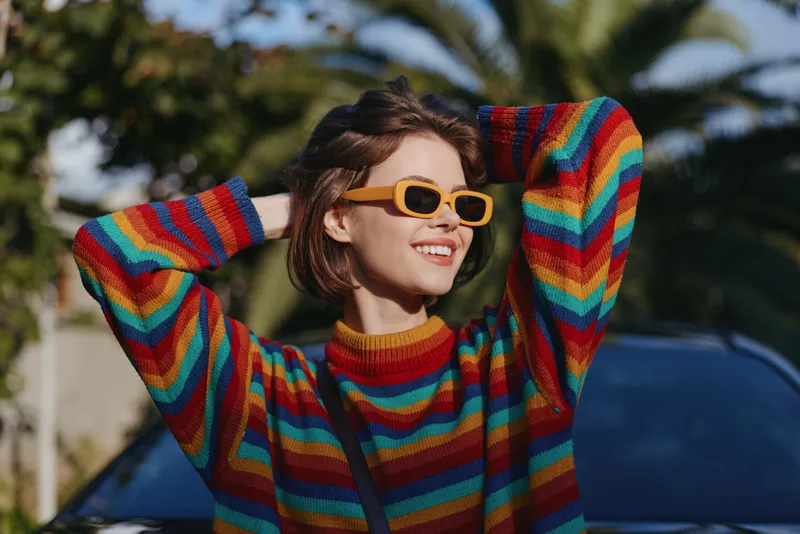 Woman smile sweater sunglasses rainbow portrait of a young woman outdoors, colorful striped sweater, short hair, casual fashion, sunny day, relaxed joyful expression near car.