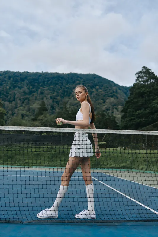 Athletic young woman in sporty outfit walking on a tennis court with a backdrop of green mountains and a cloudy sky, showcasing confidence and vitality