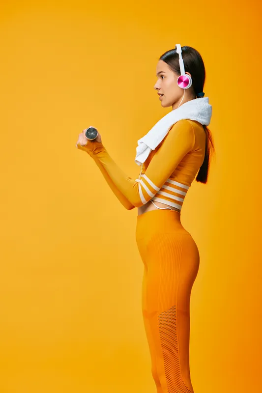 Fitness woman in orange athleticwear lifts a dumbbell against a vibrant orange backdrop, headphones on and a towel draped over shoulders, focused on workout and endurance.