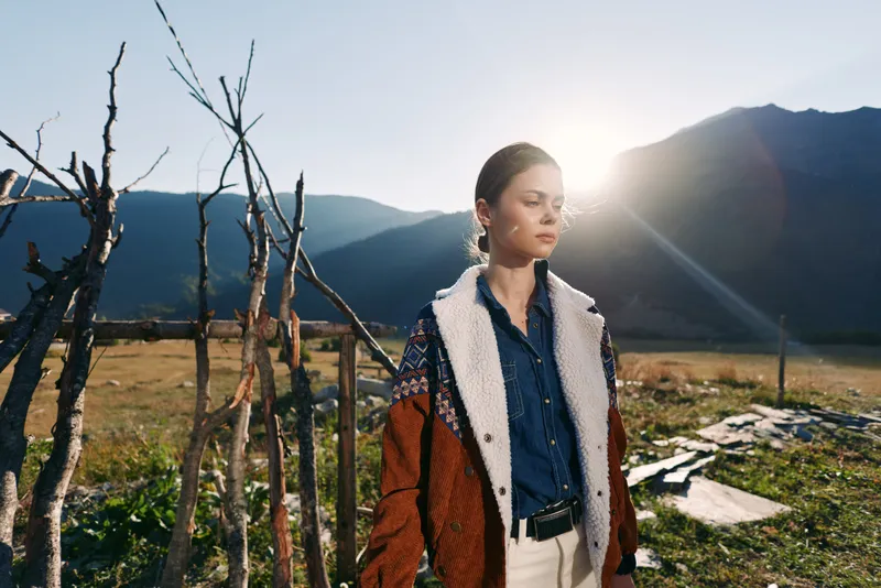Woman portrait in mountains with warm sunlight, fashion outfit and layered jacket standing outdoors by rustic wooden fence, serene landscape and travel lifestyle mood.