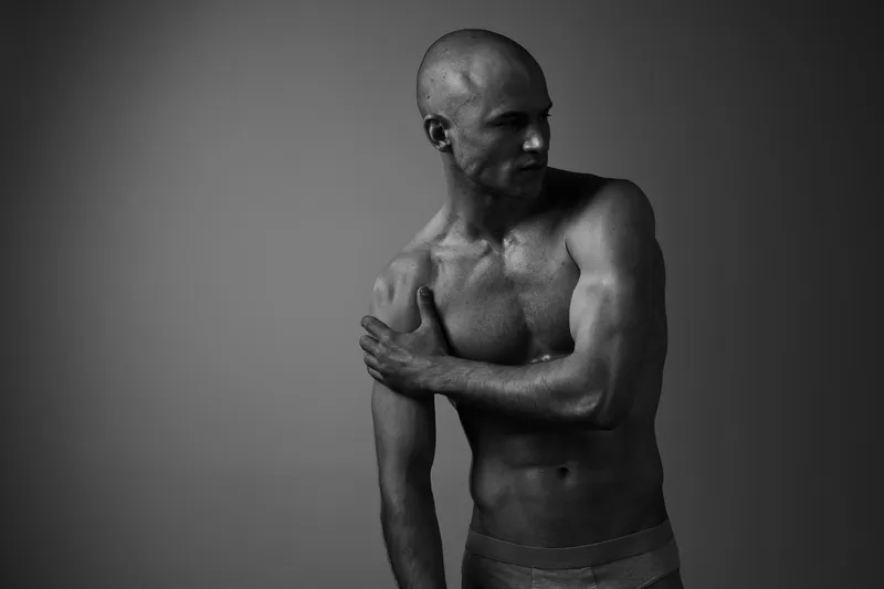 Black and white image of a man posing thoughtfully, showcasing his muscular physique and emotional expression in a dramatic lighting setting.