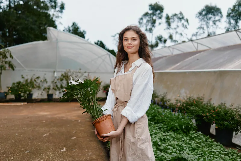 Young woman with a potted plant in a greenhouse, wearing a beige apron and white shirt, showcasing a cheerful demeanor amidst lush greenery