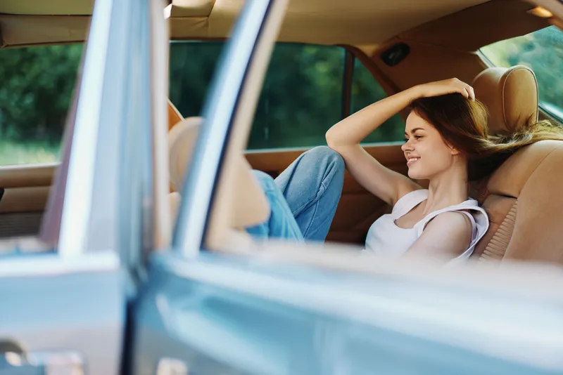 Smiling young woman relaxing in a car, wearing a white tank top and denim jeans, enjoying a sunny day outdoors, with a blurred green background.