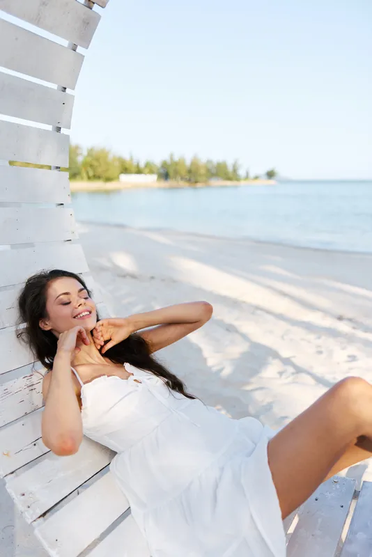 Relaxed woman in a white dress lounges against a rustic beach shelter as sunlight graces soft sand and calm water, capturing tranquil summer leisure.
