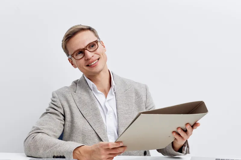 Professional businesswoman with short hair and glasses, wearing a light gray blazer and white shirt, smiling and holding a folder, in a bright office setting. Corporate, professional, confident, work,