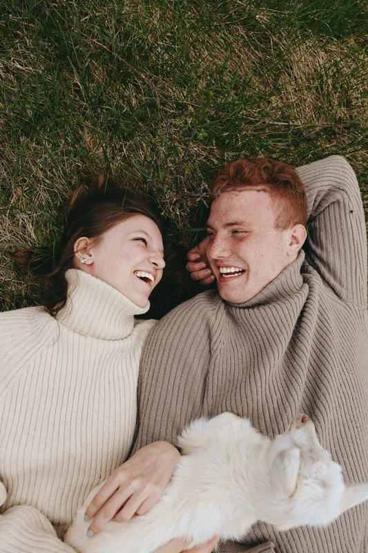 A couple lies on the grass in knit sweaters, smiling and cuddling with a fluffy pet, creating a cozy lifestyle moment outdoors that expresses affection, warmth, and togetherness.