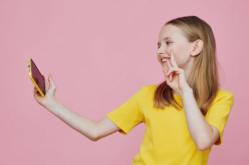 Happy girl in a yellow t shirt taking a selfie with a smartphone against a pink background, showcasing joy and modern lifestyle
