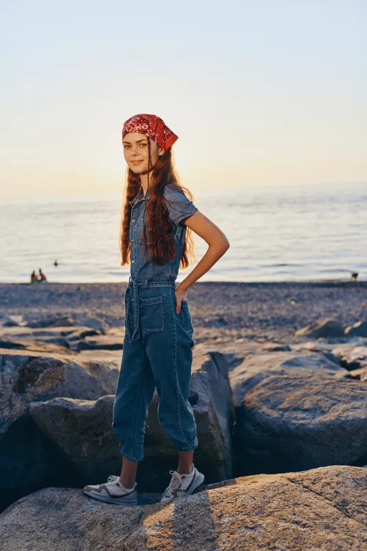 lifestyle woman in modern boho-western denim street style with a bandana standing near rocks by the sea in warm film color tones