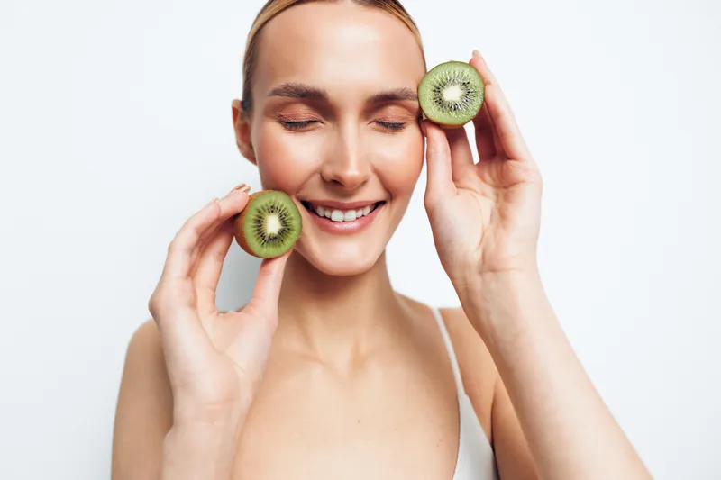 A smiling young woman with clear skin holds two halves of a kiwi fruit near her face. The bright studio background highlights natural beauty and healthy lifestyle concept.