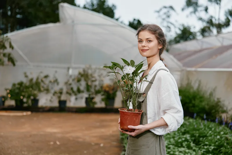 Young woman holding a potted plant in a greenhouse, showcasing a serene smile and a connection with nature, dressed in casual, earth toned clothing