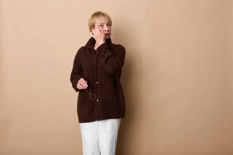 Woman in dark coat in studio, beige backdrop, hand near mouth, laughing expression, calm pose, winter fashion look, soft light, minimal background, copy space, portrait