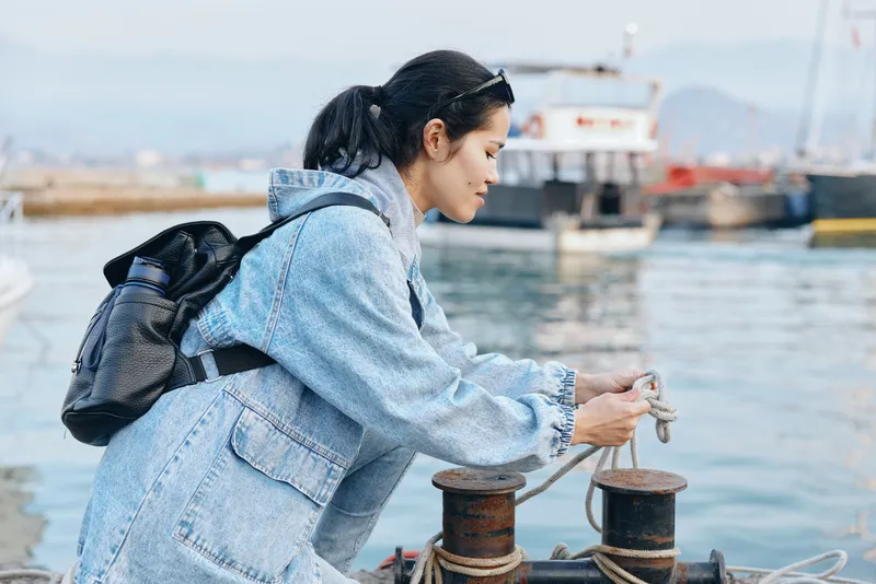 Woman in denim jacket at a harbor handles rope beside a moored boat, focused on task by the water, maritime setting, backpack ready for travel, streetwear style meets practical workwear