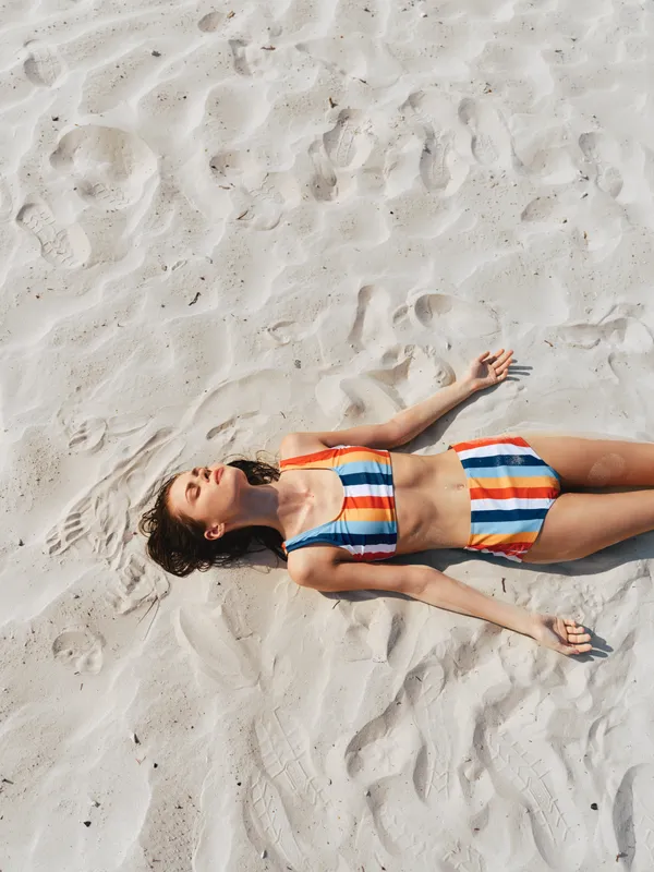 A woman lying on white sandy beach wearing a colorful striped bikini, sunlit skin and relaxed pose, conveying summer leisure and carefree vacation vibes.