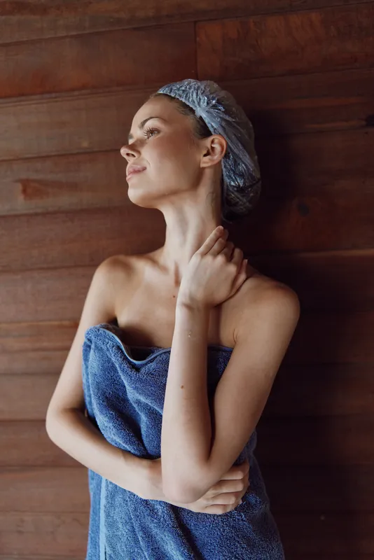 young woman wrapped in blue towel with shower cap poses against wooden wall, relaxed and fresh after bath, natural beauty and calm atmosphere indoors